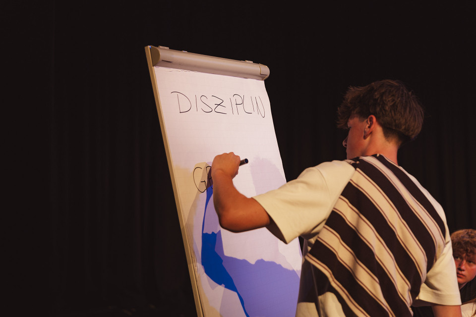 Ein junger Mann schreibt mit einem Marker auf ein Flipchart, auf dem groß das Wort „DISZIPLIN“ steht. Die Szene wirkt wie eine Präsentation oder ein Workshop auf einer Bühne vor dunklem Hintergrund.