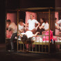Auf einer Theaterbühne spielen mehrere Darsteller in weißen Shirts eine turbulente Szene in einem metallenen Bett mit rosa Vorhängen. Spiegelwände reflektieren die Figuren, während eine Person quer über dem Bett liegt und die anderen sie dramatisch festhalten.