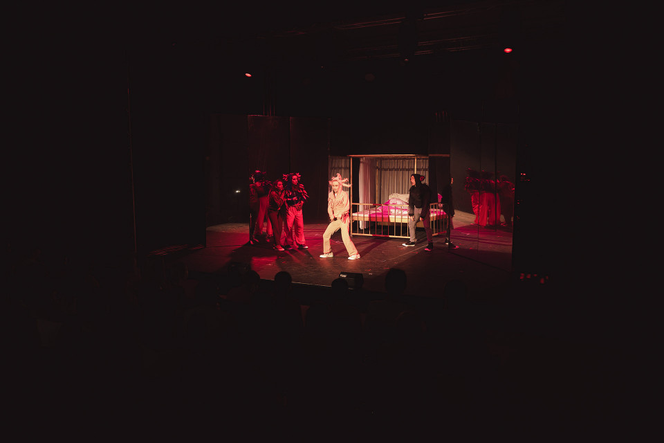 Auf einer dunklen Theaterbühne stehen mehrere Darsteller im roten Licht, während in der Mitte eine hell gekleidete Person hervorgehoben wird. Im Hintergrund ist ein Bett mit Vorhängen zu sehen, daneben reflektieren schwarze Spiegelwände die Szene.