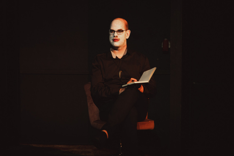 Eine kahlköpfige Person mit Brille, weiß geschminktem Gesicht und rotem Lippenstift sitzt in dunkler Kleidung auf einem Stuhl. Sie hält ein aufgeschlagenes Buch in den Händen und wird vor schwarzem Hintergrund warm beleuchtet.