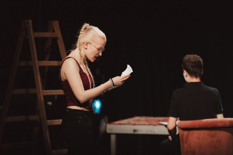 Eine blonde Frau mit geflochtenem Zopf und Brille steht auf einer dunklen Bühne und liest konzentriert von einem Blatt Papier. Vor ihr sitzt eine Person mit dem Rücken zur Kamera an einem Tisch, während im Hintergrund eine Holzleiter zu sehen ist.