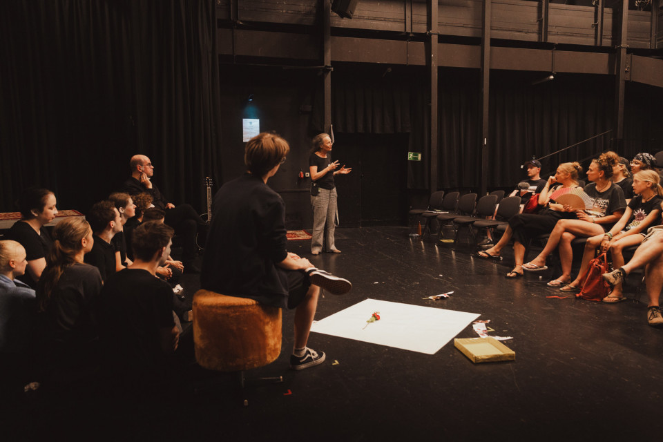 Eine Gruppe von Menschen sitzt in einem dunklen Theater- oder Proberaum im Kreis und hört einer stehenden Frau zu, die offenbar etwas erklärt. Auf dem Boden liegen einige Requisiten, während Stühle, schwarze Vorhänge und Bühnenbeleuchtung die Szene umgeben.