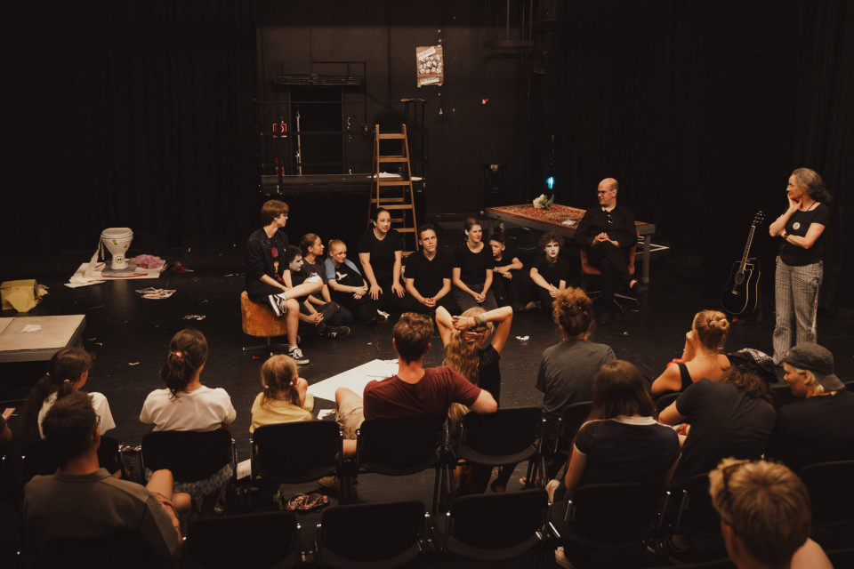 Eine Gruppe von Jugendlichen und Erwachsenen sitzt in einem dunklen Theaterraum im Kreis auf der Bühne und hört einer stehenden Frau zu. Im Hintergrund sind Requisiten wie eine Leiter, ein Teppich, eine Gitarre und Bühnenvorhänge zu sehen.