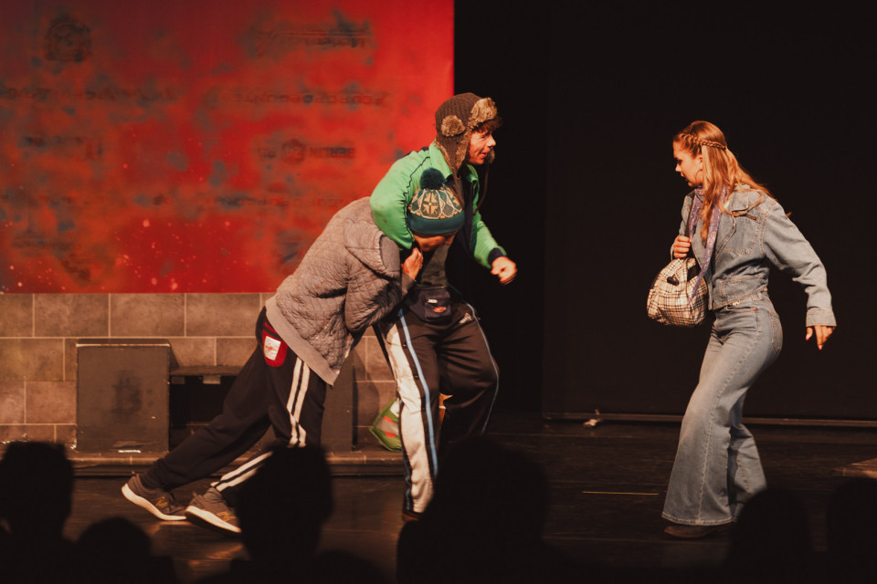 Auf einer Theaterbühne stehen drei Darsteller in einer angespannten Szene, während zwei Männer links in Bewegung sind und eine Frau rechts mit einer Tasche zurückweicht. Der Hintergrund ist dunkel mit einer rot beleuchteten Wand, und im Vordergrund sind die Silhouetten des Publikums zu sehen.