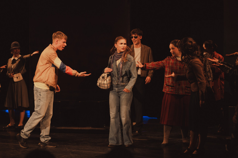 Auf einer dunkel beleuchteten Theaterbühne stehen mehrere junge Menschen in Kostümen und gestikulieren dramatisch miteinander. Im Mittelpunkt wirkt eine Frau in Jeansjacke verunsichert, während die anderen Figuren sie umringen und auf sie einreden.
