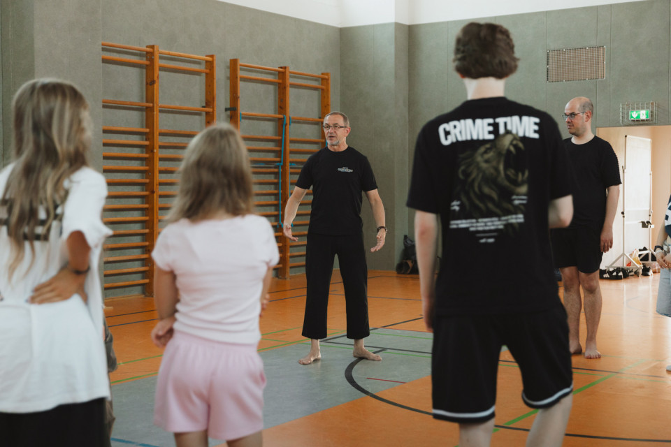 In einer Turnhalle steht ein barfüßiger Mann in schwarzer Kleidung vor einer Gruppe und scheint etwas zu erklären oder anzuleiten. Mehrere Personen hören ihm zu, während an der Wand Holz-Sprossenwände und Sportgeräte zu sehen sind.