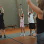 Mehrere Kinder und Jugendliche stehen in einer Sporthalle und heben bei einer Übung die Arme nach oben. Im Vordergrund ist eine Person unscharf zu sehen, während im Hintergrund Bänke und Hallenmarkierungen sichtbar sind.