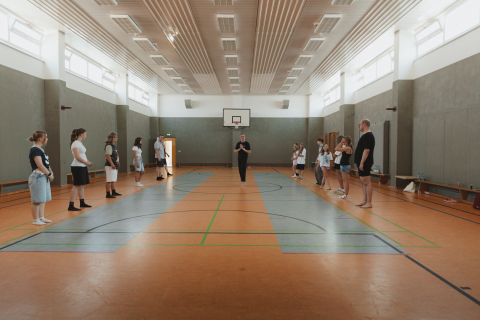 In einer hellen Sporthalle stehen mehrere Personen in zwei Reihen einander gegenüber, während eine Person in der Mitte offenbar etwas erklärt. Im Hintergrund sind ein Basketballkorb, Turnbänke und hohe Fenster zu sehen.