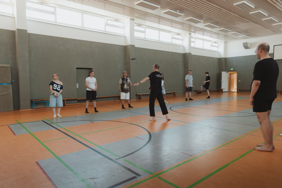 In einer großen Sporthalle stehen mehrere Jugendliche und zwei Erwachsene barfuß verteilt auf dem Spielfeld. Eine Person in der Mitte scheint die Gruppe anzuleiten oder eine Übung zu erklären.