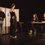 Auf einer dunklen Bühne stehen zwei Personen vor einer Stellwand mit Plakaten, während zwei junge Frauen an einem Tisch sitzen und ihnen gegenüberblicken. Die Szene wirkt wie eine Beratung, Prüfung oder Theaterprobe mit schlichtem Bühnenaufbau und gedämpftem Licht.