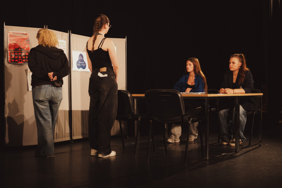 Auf einer dunklen Bühne stehen zwei Personen vor einer Stellwand mit Plakaten, während zwei junge Frauen an einem Tisch sitzen und ihnen gegenüberblicken. Die Szene wirkt wie eine Beratung, Prüfung oder Theaterprobe mit schlichtem Bühnenaufbau und gedämpftem Licht.