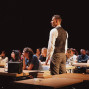 Ein Mann in Hemd und Weste steht in einem abgedunkelten Raum zwischen Reihen von Tischen, an denen viele junge Menschen sitzen. Die Szene wirkt wie eine Unterrichts- oder Theatervorstellung, mit Büchern und einer Tasche auf den Tischen im Vordergrund.