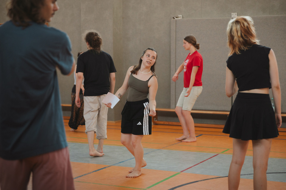 Mehrere junge Menschen bewegen sich barfuß in einer Turnhalle, als würden sie an einer Tanz- oder Theaterübung teilnehmen. In der Mitte steht eine Person mit einem Blatt Papier in der Hand und blickt zu den anderen Teilnehmenden.