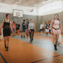 In einer hellen Sporthalle mit Basketballkorb laufen mehrere junge Menschen locker umher, teils barfuß oder in Socken. Im Vordergrund gehen drei Personen in auffälliger Freizeitkleidung auf die Kamera zu, während im Hintergrund weitere Gruppen miteinander interagieren.