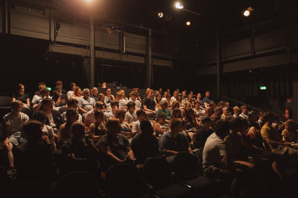 Ein großes Publikum sitzt in einem dunklen Theater- oder Veranstaltungsraum auf ansteigenden Sitzreihen. Warme Bühnenlichter beleuchten Teile der Zuschauer, während der restliche Raum überwiegend im Schatten liegt.