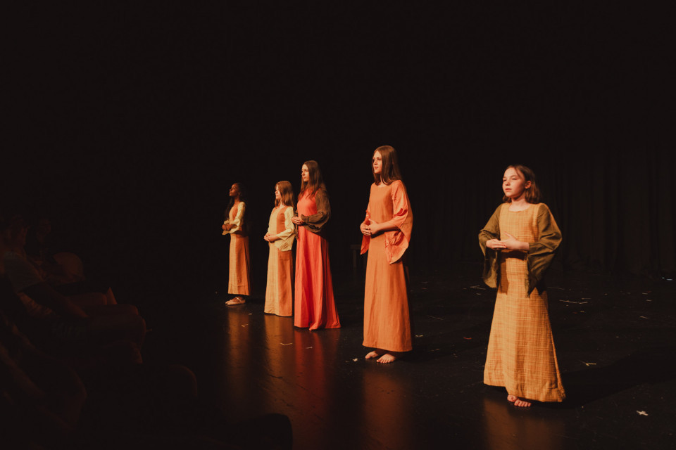 Fünf Mädchen stehen in langen, warmfarbenen Kostümen auf einer dunkel beleuchteten Theaterbühne. Links sind schemenhaft Zuschauer zu erkennen, während die Darstellerinnen ruhig mit gefalteten Händen nach vorn blicken.