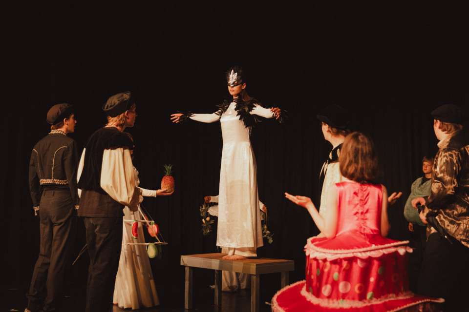 Auf einer dunklen Theaterbühne steht eine Person in einem weißen Kostüm mit Maske auf einem Tisch, während andere kostümierte Darsteller sie umringen. Einige Figuren tragen auffällige Requisiten wie eine Ananas und ein großes rosa Kuchenkostüm, wodurch die Szene fantasievoll und inszeniert wirkt.