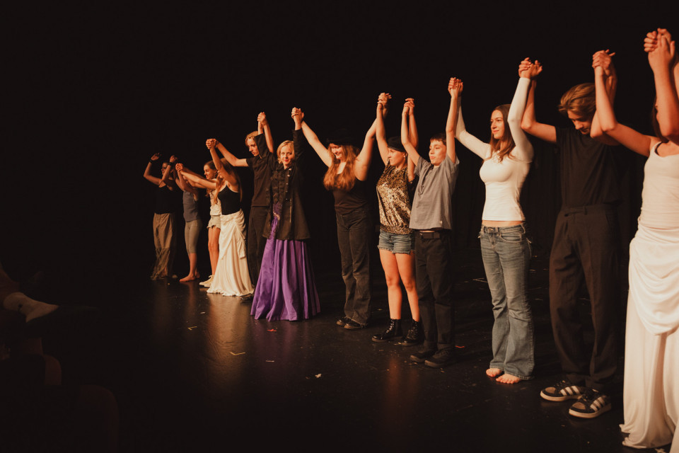 Eine Gruppe von Darstellerinnen und Darstellern steht auf einer dunklen Theaterbühne in einer Reihe und hält sich mit erhobenen Armen an den Händen. Warmes Bühnenlicht beleuchtet die unterschiedlich gekleideten Personen, als würden sie sich nach einer Aufführung verbeugen oder vom Publikum feiern lassen.