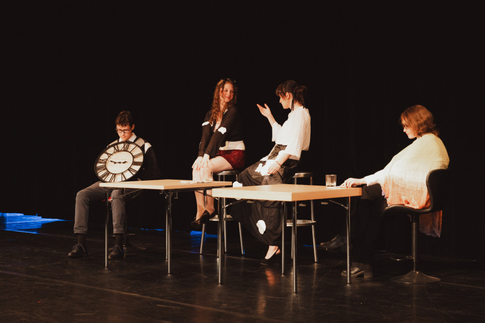 Auf einer dunklen Theaterbühne sitzen vier Personen an zwei Tischen und wirken wie in einer Szene oder Aufführung. Links hält ein Mann eine große Uhr-Requisite, während die anderen drei Personen auf Hockern sitzen und miteinander interagieren.