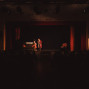 Auf einer dunkel beleuchteten Theaterbühne stehen zwei Schauspieler im Scheinwerferlicht vor einem roten Vorhang. Im Vordergrund sind die Zuschauer nur als dunkle Silhouetten zu erkennen, während auf der Bühne ein Sofa und ein Sessel als Kulisse stehen.