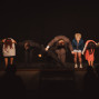Eine Gruppe von Schauspielerinnen und Schauspielern verbeugt sich Hand in Hand auf einer dunkel beleuchteten Bühne. Im Vordergrund sind die Silhouetten des Publikums zu sehen, das der Aufführung zuschaut.