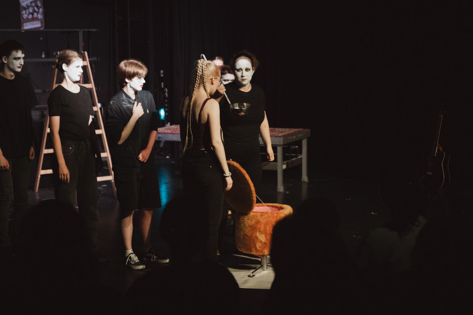 Auf einer dunklen Theaterbühne stehen mehrere schwarz gekleidete Personen mit weiß geschminkten Gesichtern im Scheinwerferlicht. Im Vordergrund sind Publikumssilhouetten zu sehen, während auf der Bühne ein orangefarbener Sessel, eine Leiter und eine Gitarre im Hintergrund stehen.