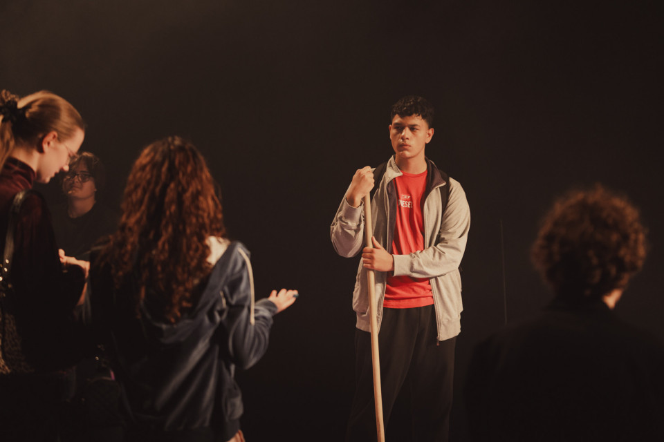 Ein junger Mann steht auf einer dunklen Bühne und hält einen langen Holzstab, während er ernst in Richtung einer Gruppe blickt. Um ihn herum stehen mehrere Personen im Halbdunkel, wodurch die Szene wie eine Theaterprobe wirkt.