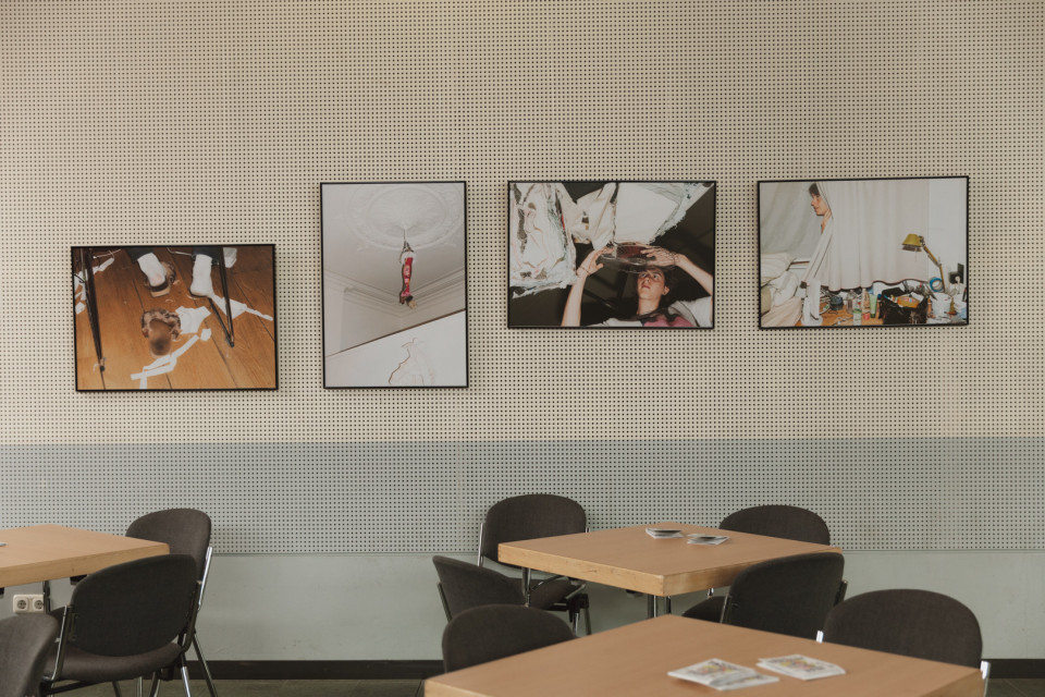 An einer perforierten Wand hängen vier gerahmte Fotografien mit ungewöhnlichen Innenraumszenen. Davor stehen mehrere Holztische und dunkle Stühle in einem ruhigen Ausstellungs- oder Aufenthaltsraum.