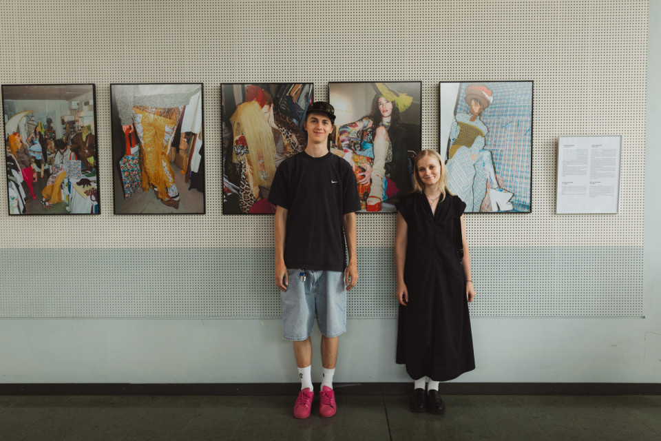Zwei Personen stehen in einer Galerie vor einer perforierten Wand mit mehreren gerahmten Mode- und Kunstfotografien. Links trägt die Person ein schwarzes T-Shirt, Jeansshorts und pinke Schuhe, rechts steht eine Person in einem langen schwarzen Kleid.