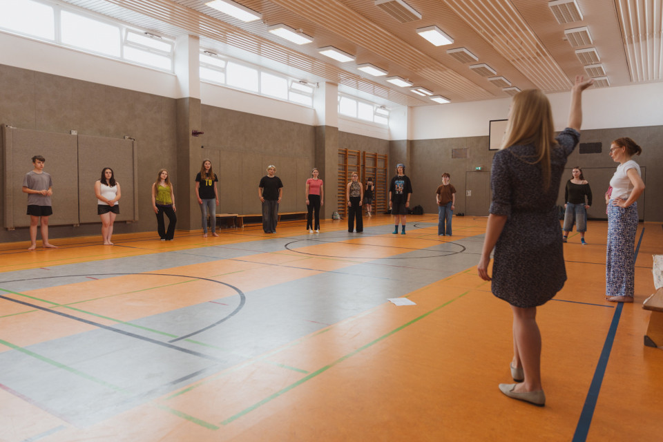 In einer Turnhalle stehen mehrere Personen weit verteilt im Kreis und schauen zu einer Frau, die vorne mit erhobenem Arm Anweisungen gibt. Der Raum ist hell, mit Sportlinien auf dem Boden, hohen Fenstern und einer Holzdecke ausgestattet.