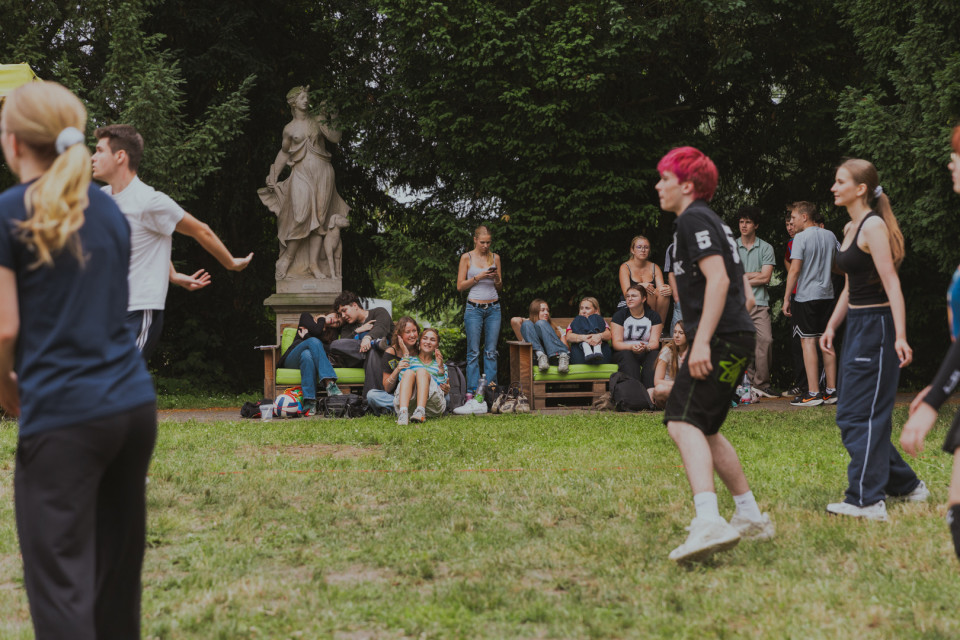Eine Gruppe junger Menschen verbringt Zeit auf einer Wiese in einem Park, einige spielen offenbar Volleyball, während andere auf Bänken sitzen oder zuschauen. Im Hintergrund stehen dichte Bäume und eine klassische Statue neben den Sitzgelegenheiten.