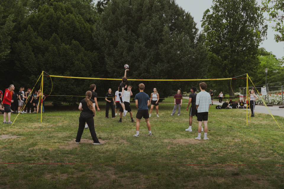 Eine Gruppe Jugendlicher spielt auf einer Wiese im Park Volleyball an einem gelben Netz. Im Hintergrund stehen Bäume und weitere Zuschauer sitzen oder stehen am Spielfeldrand.