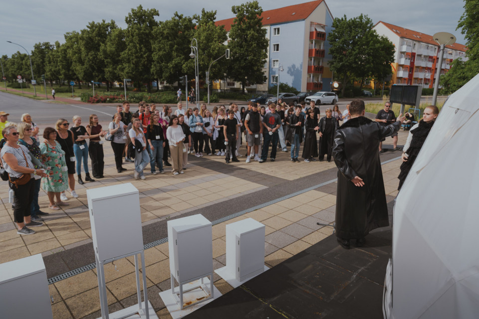 Eine größere Gruppe Menschen versammelt sich auf einem Platz vor einer kleinen Bühne und hört einer Person in einem langen schwarzen Mantel zu. Im Hintergrund sind Wohnhäuser, Bäume und eine Straße zu sehen, während auf der Bühne weiße Kästen und ein großes weißes Zeltelement stehen.