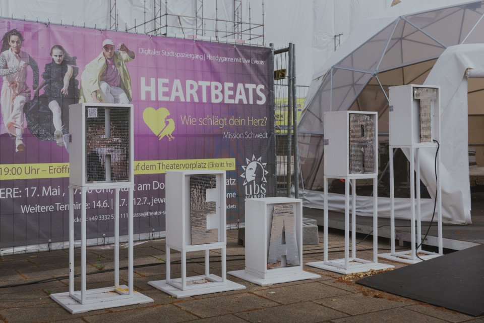 Vor einem großen violetten Werbebanner für „HEARTBEATS“ stehen weiße, beleuchtete Buchstabeninstallationen, die „HEART“ bilden. Rechts daneben befindet sich eine transparente Zelt- oder Bühnenkonstruktion auf einem gepflasterten Platz.