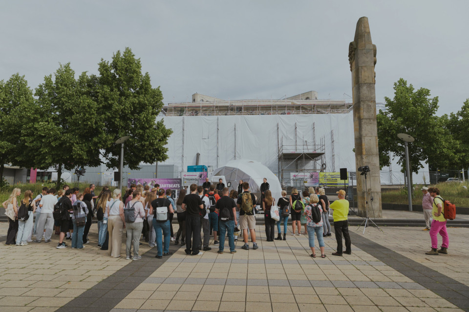 Eine Gruppe von Menschen versammelt sich auf einem gepflasterten Platz vor einer kleinen Bühne oder Kuppel, während im Hintergrund ein großes eingerüstetes Gebäude verhüllt ist. Rechts steht ein hohes steinernes Denkmal zwischen Bäumen, und die Szene wirkt wie eine öffentliche Veranstaltung im Freien.