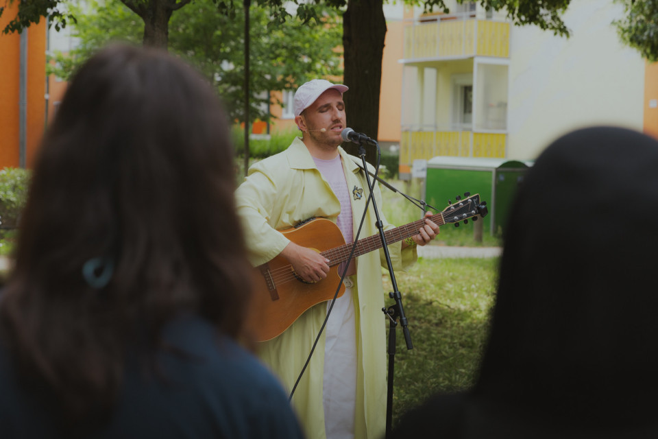 Ein Musiker in heller Kleidung und weißer Kappe singt im Freien in ein Mikrofon und spielt dabei Gitarre. Im Vordergrund sind unscharf die Köpfe von Zuschauern zu sehen, während im Hintergrund Bäume und Wohnhäuser stehen.