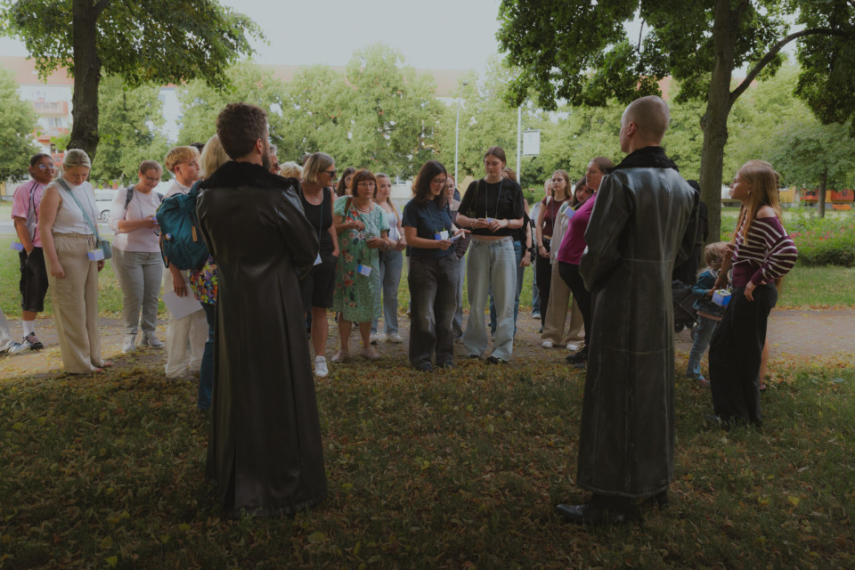 Eine Gruppe von Menschen steht in einem schattigen Park unter Bäumen und hört zwei Personen in langen schwarzen Mänteln zu. Einige Teilnehmende tragen Namensschilder und halten Zettel, während im Hintergrund Grünflächen und Wohngebäude zu sehen sind.