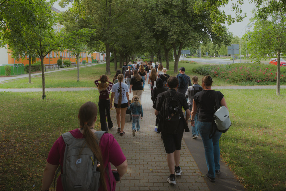 Eine größere Gruppe von Menschen geht auf einem gepflasterten Weg durch eine grüne, von Bäumen gesäumte Umgebung. Links stehen Wohngebäude, rechts verlaufen eine Straße und Blumenbeete entlang der Wiese.
