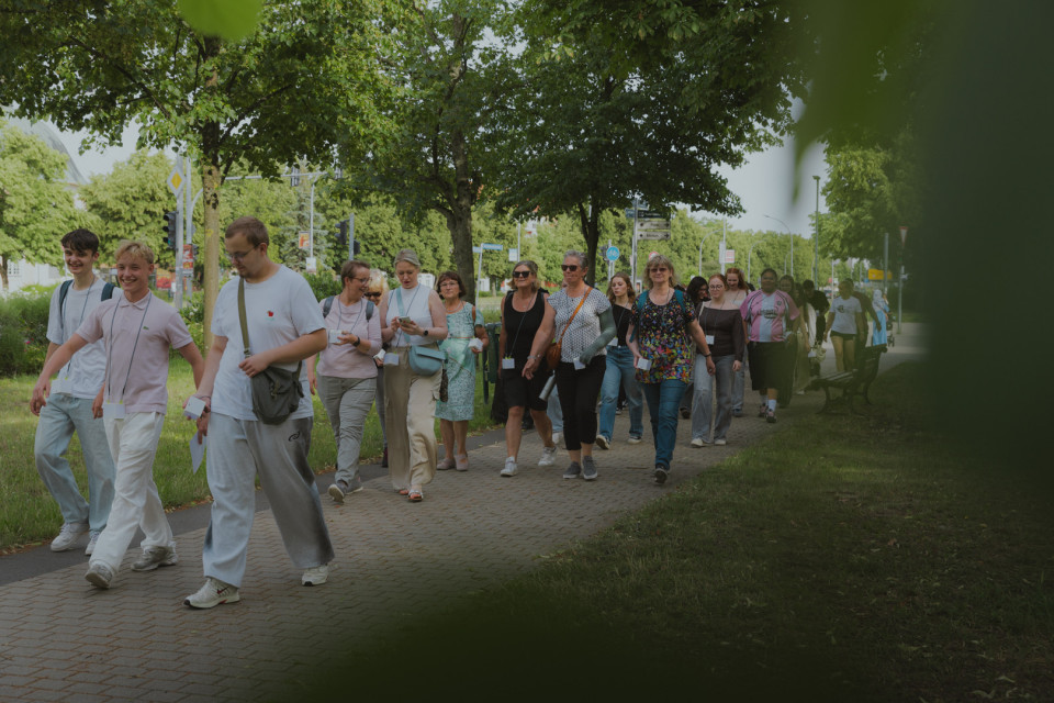 Eine größere Gruppe von Menschen geht an einem sonnigen Tag auf einem von Bäumen gesäumten Gehweg entlang. Viele tragen Namensschilder und wirken wie Teil einer gemeinsamen Führung oder Veranstaltung.