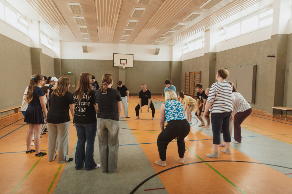 Eine Gruppe von Menschen steht in einer Sporthalle im Kreis und scheint gemeinsam eine Übung oder ein Spiel zu machen. Im Hintergrund sind ein Basketballkorb, Turngeräte und hohe Fenster zu sehen.