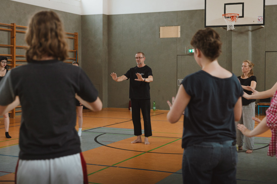 In einer Sporthalle leitet ein barfüßiger Mann in schwarzer Kleidung eine Gruppe bei ruhigen Bewegungsübungen an. Mehrere Teilnehmende stehen im Kreis und folgen seinen Handbewegungen, während im Hintergrund ein Basketballkorb zu sehen ist.
