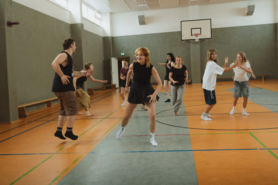 Mehrere junge Erwachsene bewegen sich lachend und spielerisch in einer Turnhalle mit Basketballkorb. Sie tragen Freizeit- oder Sportkleidung und scheinen gemeinsam zu tanzen oder eine Übung zu machen.