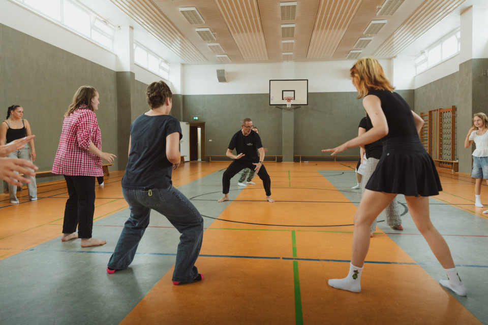 Eine Gruppe von Menschen steht barfuß oder in Socken in einer Sporthalle und macht gemeinsam Bewegungsübungen. In der Mitte leitet ein Mann die Übung an, während die Teilnehmenden im Kreis um ihn herum seine Haltung nachahmen.