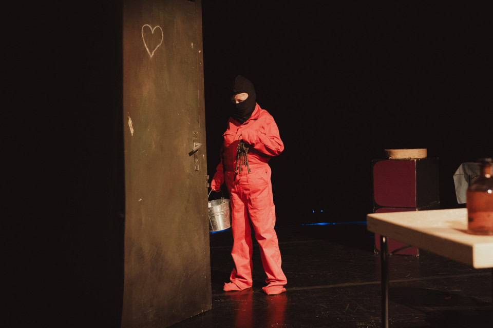 Eine Person in rotem Overall und schwarzer Maske steht auf einer dunklen Bühne neben einer hohen Tür, auf die ein Herz gezeichnet ist. Sie hält einen Schlüsselbund und einen Metalleimer, während rechts im Bild ein Tisch und Requisiten schwach beleuchtet sind.