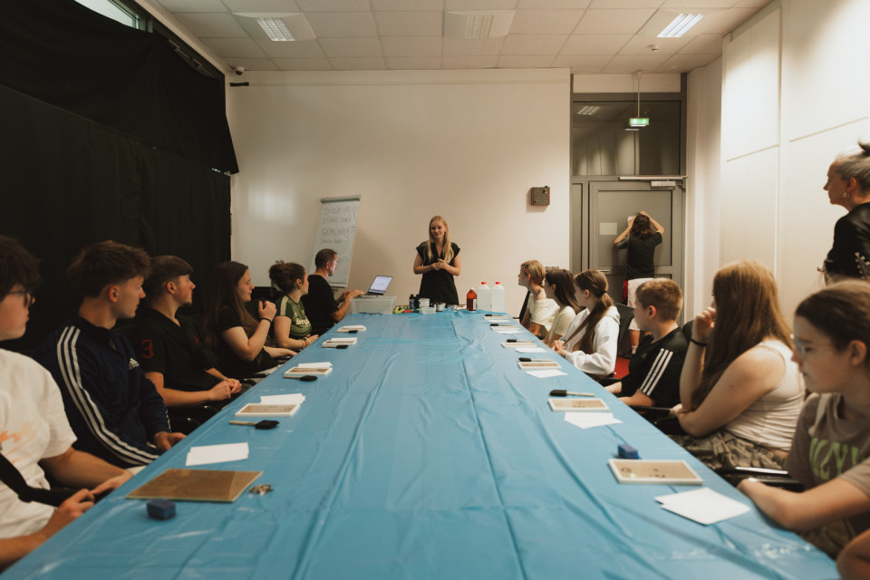 Eine Gruppe Jugendlicher sitzt in einem Seminarraum an einem langen Tisch mit blauer Abdeckung und schaut einer stehenden Kursleiterin zu. Auf dem Tisch liegen Papier, kleine Materialien und Geräte, während im Hintergrund ein Flipchart und weitere Personen zu sehen sind.