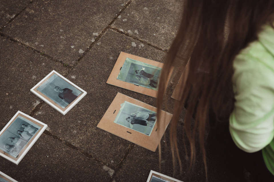 Eine Person mit langen Haaren betrachtet mehrere gerahmte und auf Karton liegende alte Fotografien auf einem steinernen Boden. Die Bilder wirken verblasst und zeigen Porträts sowie Gruppenaufnahmen in einem nostalgischen Stil.