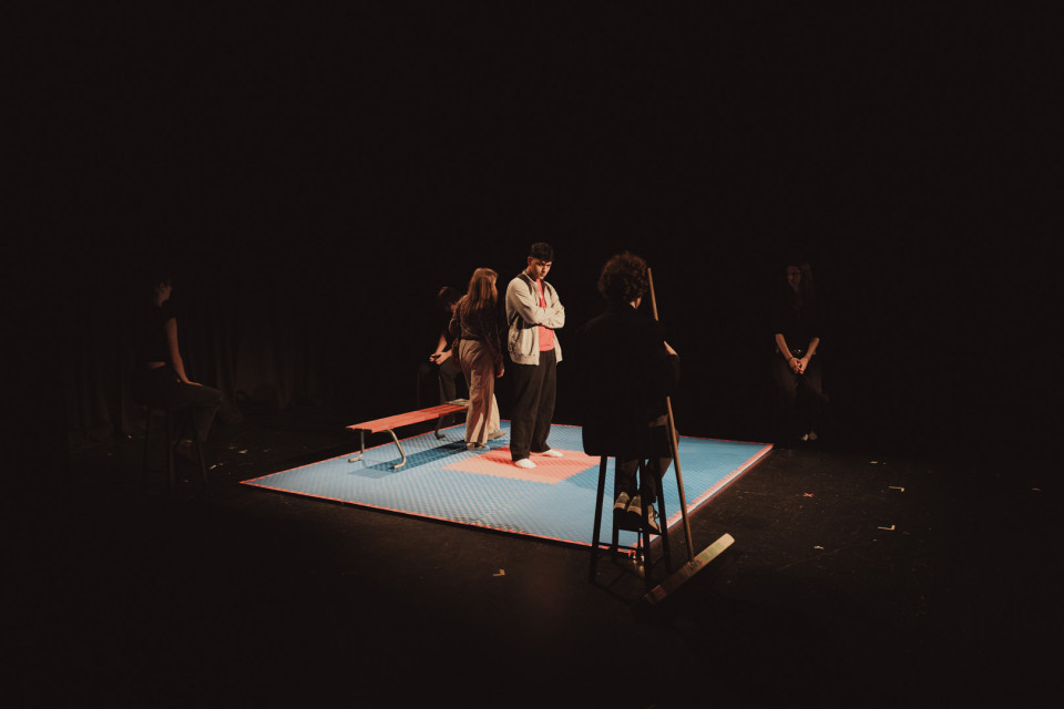 Auf einer dunklen Theaterbühne stehen mehrere Personen um eine hell beleuchtete, blau-rote Matte mit einer Bank in der Mitte. Die Szene wirkt wie eine Probe oder Aufführung, bei der die Figuren verteilt im Raum stehen und im Scheinwerferlicht unterschiedliche Positionen einnehmen.