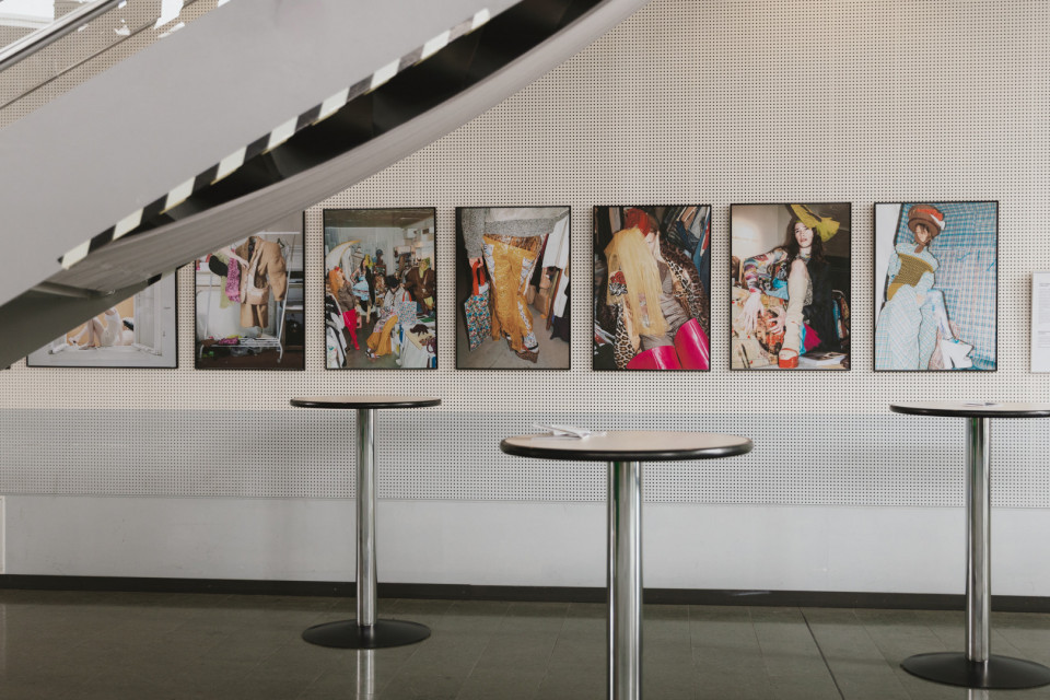 An einer hellen Wand hängen mehrere gerahmte Fotografien mit farbenfrohen Mode- und Kostümszenen. Im Vordergrund stehen runde Stehtische, während links oben eine Treppe schräg durch das Bild verläuft.