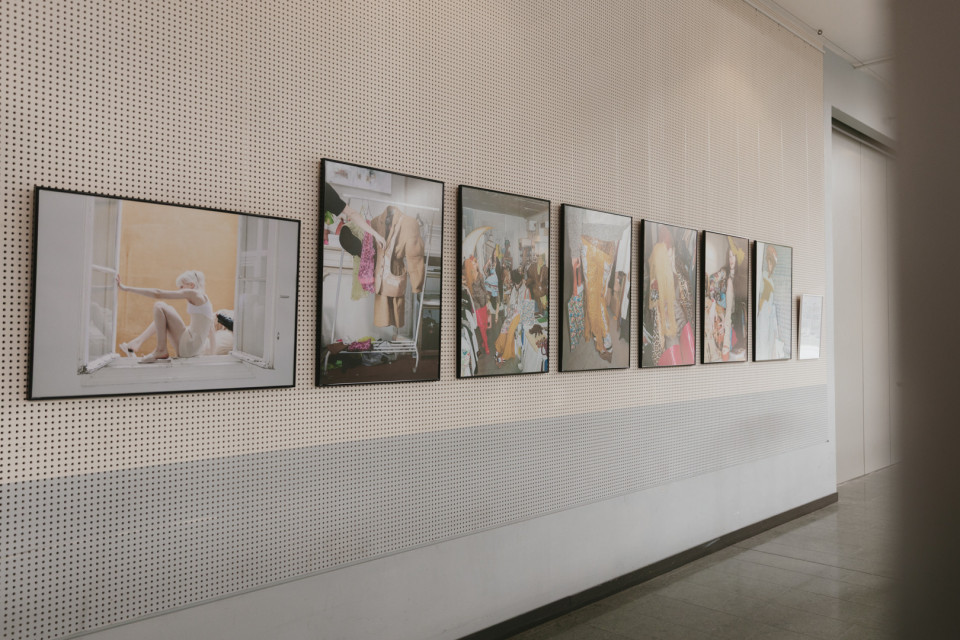 An einer langen, perforierten Galeriewand hängen mehrere gerahmte Fotografien in einer Reihe. Der helle Ausstellungsraum wirkt ruhig, mit Fliesenboden und einer Tür am Ende des Flurs.