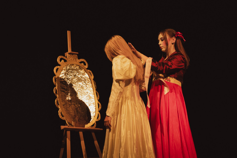 Auf einer dunklen Bühne stehen zwei Frauen in historischen Kleidern neben einem goldenen Spiegel auf einer Staffelei. Die Frau im roten Kleid berührt behutsam die Haare der Frau im hellen Kleid, während warmes Licht die Szene beleuchtet.
